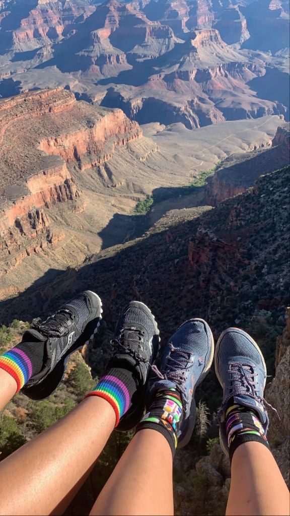 Photo taken by the author. Depicts two pairs of feet in brightly colored socks hanging over the edge of the Grand Canyon. 